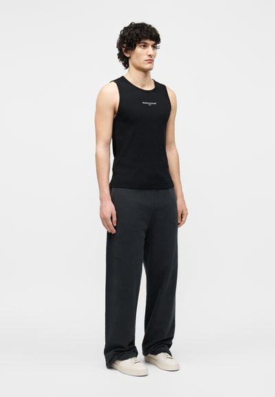 Jeune homme portant un haut noir sans manches avec un logo blanc, un pantalon ample foncé et des baskets blanches, debout sur un fond clair uni.