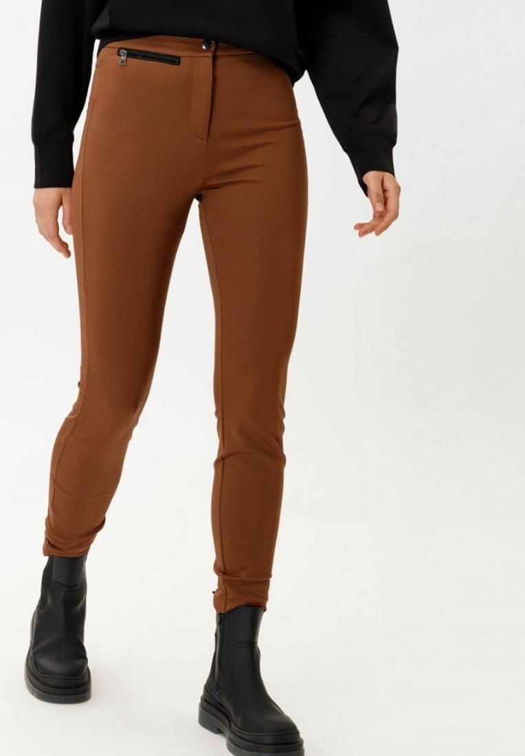 Pantalon marron ajusté en matière extensible, avec une poche zippée et des lignes épurées. Associé à des bottines noires.