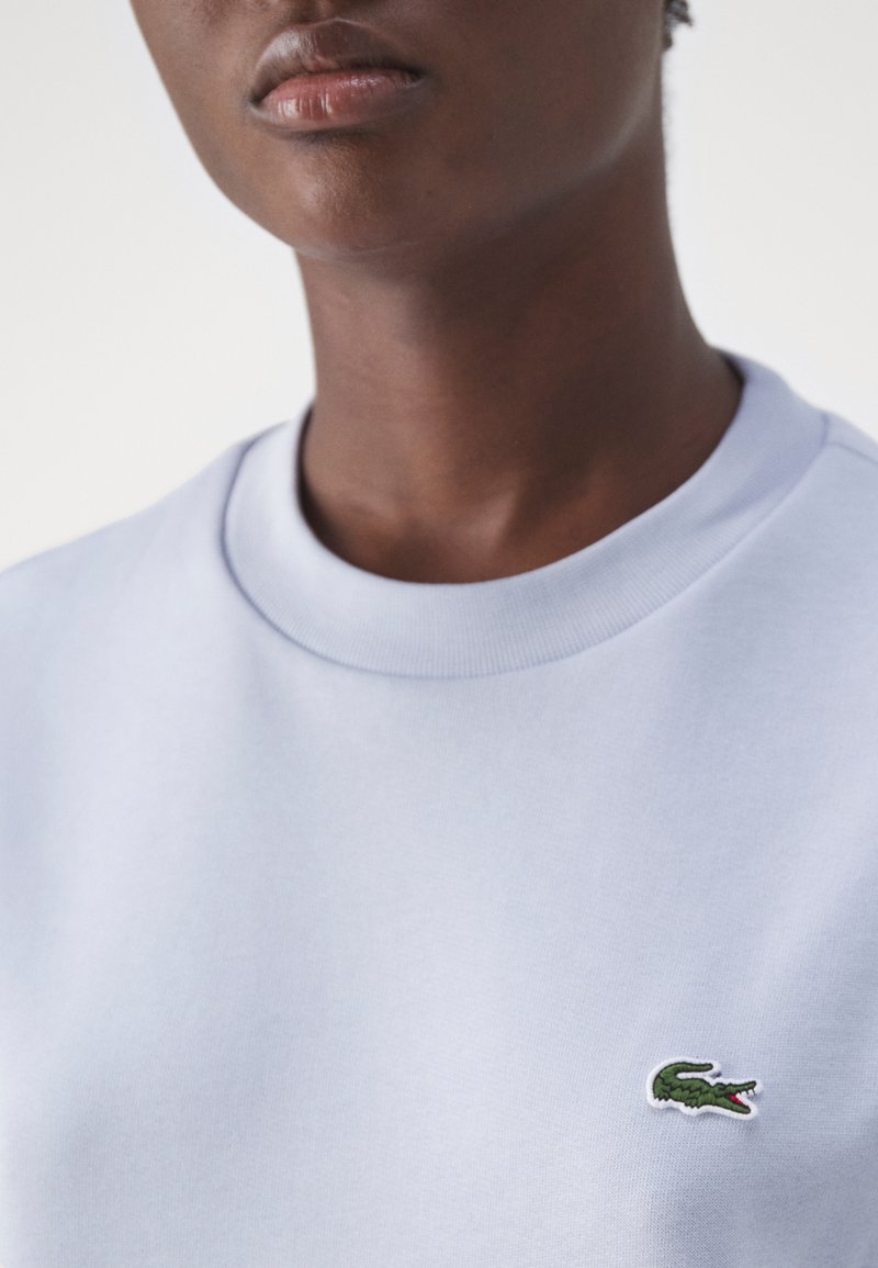 Lichtblauw sweatshirt met een gladde textuur, ronde halslijn en een klein groen krokodillenlogo op de borst.