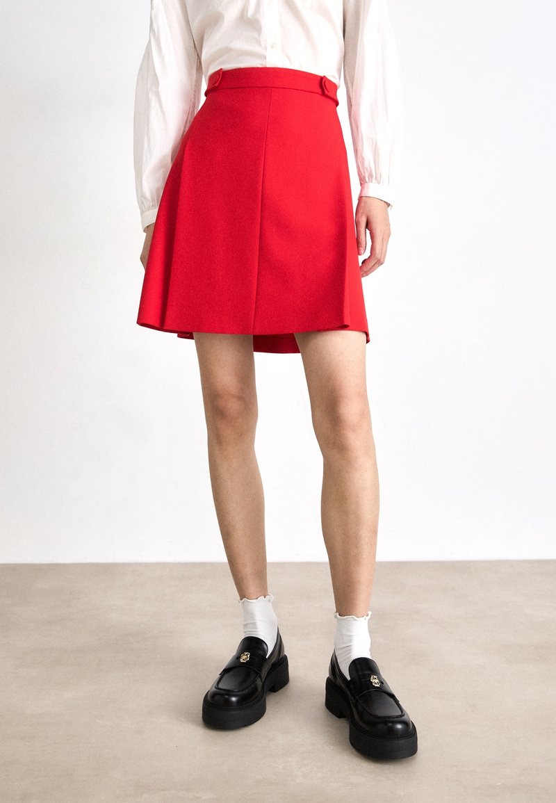 Jupe rouge en ligne A avec une taille ajustée, ornée de deux boutons. Le mannequin porte des mocassins noirs à semelles épaisses et des chaussettes blanches.