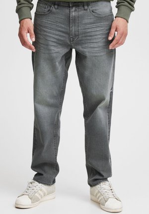 Homme portant un jean gris délavé et des baskets blanches, debout devant un fond uni avec les mains relâchées le long du corps.