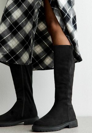 Botas de ante negras hasta la rodilla con suela plana y acabado texturizado, combinadas con una prenda de cuadros en blanco y negro que presenta una abertura lateral.