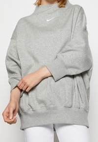 Grå sweatshirt med hög krage, oversized passform och ribbade ärmslut. Har en liten vit Nike-logga på övre vänstra sidan.