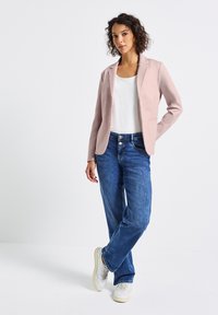 Blazer rose clair sur un top blanc, associé à un jean bleu et des baskets blanches. Le blazer possède des revers crantés et une texture lisse.