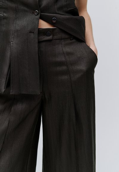 Gros plan sur un pantalon noir taille haute et un gilet sans manches assorti à motif chevrons, avec une main dans la poche, sur un fond uni.