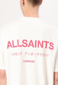 T-shirt en coton blanc avec un texte rose vif au dos, "ALLSAINTS" et "LONDRES", avec des caractères supplémentaires non latins. Manches courtes.