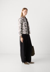 Giacca bomber stampata con uno sfondo grigio chiaro che presenta forme astratte nere e marroni, abbinata a pantaloni neri a gamba larga e scarpe a punta.
