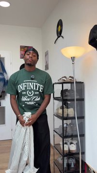 Un tânăr îmbrăcat cu un durag și o bluză verde ține o țesătură cu dungi într-o cameră care are un suport pentru pantofi, o lampă de podea și decoruri pe perete.
