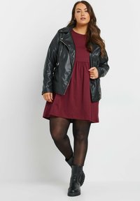 Mustapasta nahkatakki burgundinvärisen mekon päällä, polvipitkä ja ylhäältä tiukka. Mustat sukkahousut ja jykevät mustat saappaat täydentävät asukokonaisuuden.