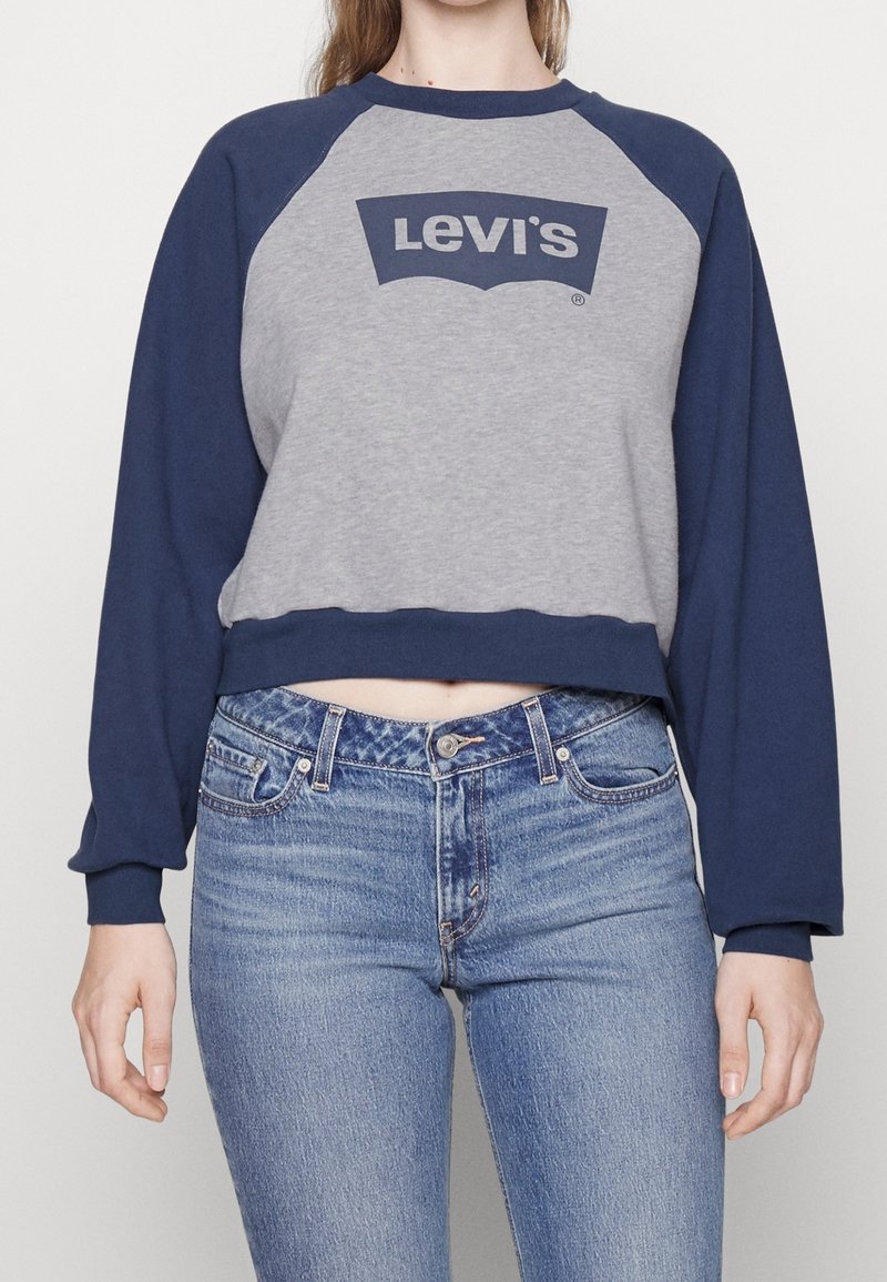 Sudadera gris corta con mangas raglán azul marino y gran logo negro de Levi's. Combinada con jeans azules descoloridos, que presentan un botón y cremallera.