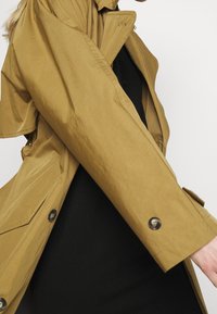 Písečný lehký trench coat se uvolněným střihem, velkými kapsami, knoflíkovými detaily a lehkým leskem, vržený přes černý top.