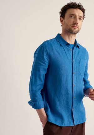 Homme aux cheveux courts et bouclés portant une chemise bleue à manches longues retroussées et un pantalon marron sur fond beige.