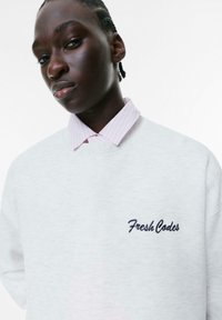 Grå sweatshirt med blød tekstur, der har "Fresh Codes" broderet i marineblå. Laget over en lyserød skjorte med krave og fine striber.