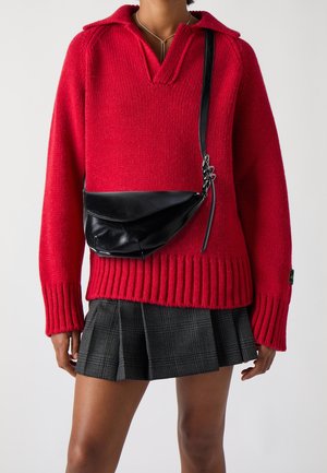 Henkilö pukeutunut punaiseen neulepuseroon, mustaan olkalaukkuun ja harmaaseen ruudulliseen laskoshameeseen.