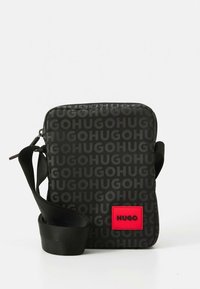 Kis fekete oldaltáska ismétlődő "HUGO" mintával és piros folttal, amelyen a "HUGO" logó látható, állítható fekete vállpánttal.