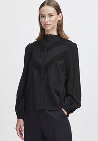 Svart långärmad blus med hög krage, som har ett texturerat chevronmönster och knäppning, tillverkad av lätt och slätt material.