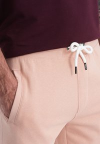 Pantalon de jogging rose clair avec poches latérales, taille élastique et cordon blanc. Le tissu semble doux et légèrement texturé.