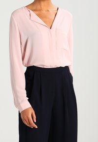 Blusa rosa chiaro con scollatura a V, maniche lunghe e tasca frontale, abbinata a pantaloni blu navy a vita alta con pieghe.