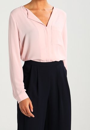 Blusa rosa chiaro con scollatura a V, maniche lunghe e tasca frontale, abbinata a pantaloni blu navy a vita alta con pieghe.
