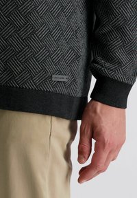 Schwarzer Pullover mit gemustertem, strukturiertem geometrischem Design, ausgestattet mit einem sichtbaren Logo-Tag und gerippten Bündchen. Kombiniert mit hellen Hosen.