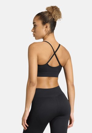 Frau mit lockigem Haar zu einem Dutt gebunden, die einen schwarzen Sport-BH mit gekreuzten Trägern und hoch taillierte Leggings trägt, von hinten vor hellem Hintergrund gezeigt.