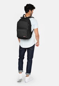 Black backpack with a zippered front pocket and logo patch, worn by a person. The material appears durable with adjustable straps and a minimalist design.