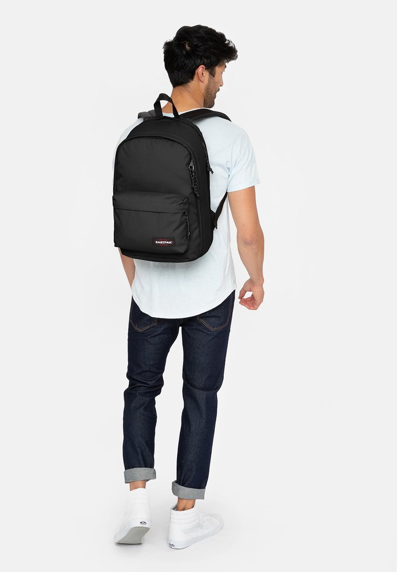 Black backpack with a zippered front pocket and logo patch, worn by a person. The material appears durable with adjustable straps and a minimalist design.