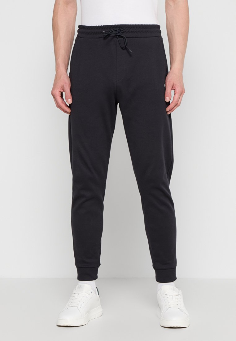 Pantaloni jogger neri in cotone con elastico in vita e cordoncino, vestibilità tapered, tasche laterali e polsini a coste. Indossati con sneakers bianche.