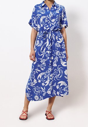 Femme portant une robe midi bleue avec des motifs floraux et feuilles blancs, manches courtes, col, ceinture à la taille et sandales rouges à brides.