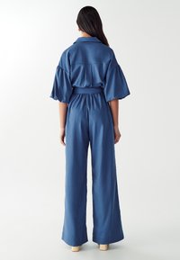 Kvinna med långt mörkt hår som bär en blå bredbent jumpsuit med ärmar i puff längd till armbågen, stående vänd bort på en vit bakgrund.