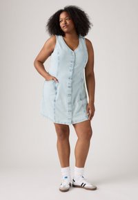 Abito senza maniche in denim blu chiaro con scollatura a V, apertura frontale con bottoni e tasche laterali. Il modello indossa sneakers bianche e calzini morbidi.