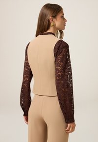 Top ajustado beige con diseño de mangas largas de encaje marrón. La vista trasera muestra una textura suave y una silueta ajustada, con detalle de botones en las mangas.