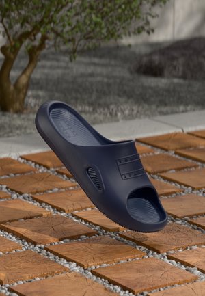 Sandalia de goma azul marino sin cordones con el logo "Tommy Jeans", flotando sobre baldosas de madera al aire libre cerca de un árbol.