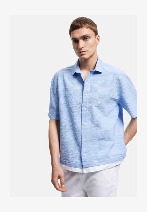 Ung mand med kort hår iført en lyseblå kortærmet ternet skjorte og hvide bukser foran en ensfarvet baggrund.