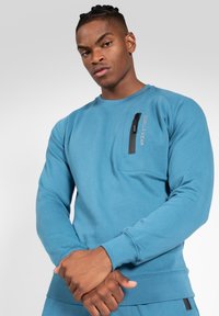 Blauwe sweatshirt met een ronde hals, lange mouwen en een ritszak op de borst. Bevat een logo gedrukt aan de zijkant en geribde manchetten.