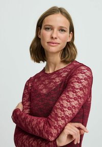 Top lungo a maniche con pizzo bordeaux, caratterizzato da un motivo floreale e scollo rotondo, con una texture trasparente e una silhouette aderente.