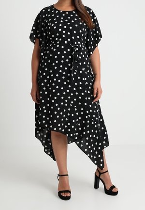 Robe asymétrique noire à pois blancs, manches courtes volantées, ceinture à la taille, portée avec des sandales noires à talons hauts et brides à la cheville.