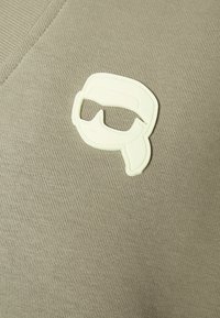 Oliwkowozielony bawełniany materiał z teksturowanej tkaniny; zawiera białą gumową naszywkę przedstawiającą parę gogli po lewej stronie.