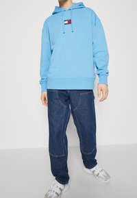 Blå huvtröja med en logotyp-applikation, tillsammans med löst sittande mörka denimjeans och vita sneakers.