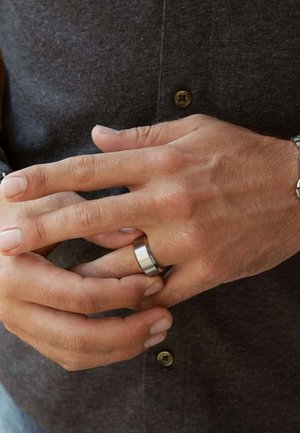 Zilveren metalen ring om een hand, met een geborstelde textuur en een gladde bovenrand. De handen zijn zachtjes samengevouwen, waardoor de vingernagels en huidskleur zichtbaar zijn.