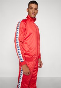 Survêtement rouge avec col montant et fermeture éclair. Présente des bandes latérales blanches contrastantes avec des motifs de logo bleus. Matière lisse et brillante.