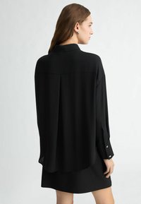 Black, long-sleeve, collared shirt with a loose fit, featuring buttoned cuffs and a back pleat detail, made from smooth fabric.