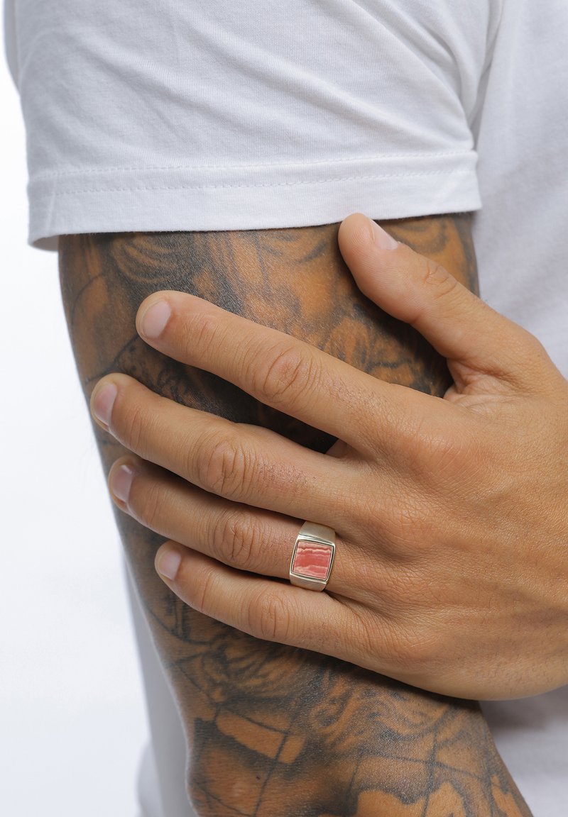 Bague en argent avec un incrustation rectangulaire de corail, tenue par une main tatouée, portant un t-shirt blanc. Texture lisse, design moderne.