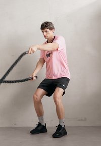 PUMA sportska odjeća sastoji se od svijetlopinkaste majice i crnih šorceva. Osoba drži crne bitke užadi u fitness pozi nasuprot neutralnoj pozadini.