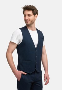 Chaleco azul marino hecho de tejido texturizado, con cuello en V, cinco botones y dos bolsillos frontales; llevado sobre una camiseta blanca.