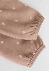 Tissu marron avec de petits motifs de cœurs roses, doté de poignets élastiques. Le matériau semble doux et texturé, adapté à un usage décontracté.