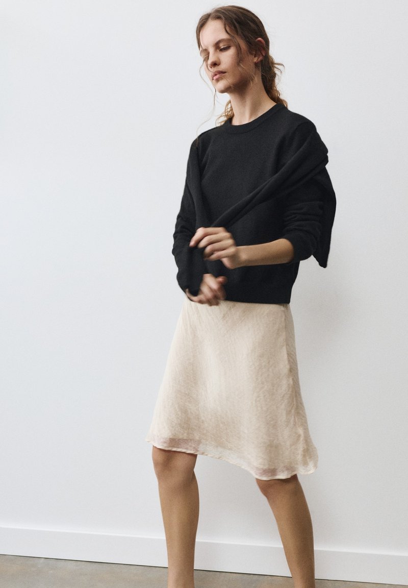 Jeune femme aux cheveux châtain clair portant un pull noir et une jupe beige, ajustant ou tirant la manche du pull, debout contre un mur blanc.