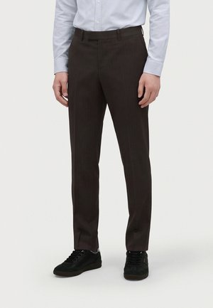 Mann trägt dunkelbraune formelle Hose, schwarze Sneaker und ein hellblaues Hemd und steht vor einem schlichten Hintergrund.