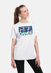Kvinde med hestehale bærer en hvid T-shirt, der viser Henri Matisse's maleri "Dansen" med teksten "DANCE" nedenfor figurerne.