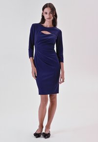 Marineblaues, tailliertes Kleid mit Dreiviertelärmeln, das über ein auffälliges Cut-out-Detail und eine drapierte Textur verfügt, kombiniert mit schwarzen Schuhen mit Schleifenakzenten.